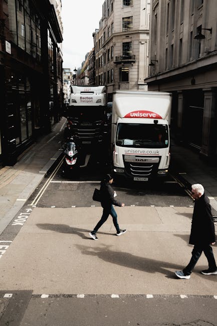 A street scene in an urban area featuring a narrow road with tall, multi-story buildings on either side. Two large moving trucks, branded with Uniserve and Becton Transport, are parked close to the cu