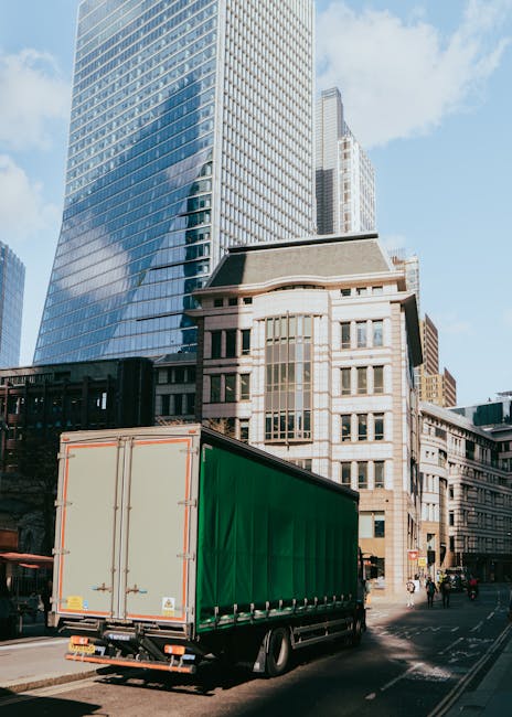 A street scene in an urban area featuring a narrow road with tall, multi-story buildings on either side. Two large moving trucks, branded with Uniserve and Becton Transport, are parked close to the cu