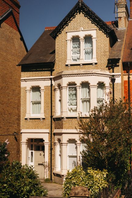 A row of mid-20th-century brick terraced houses with pitched roofs and bay windows line a busy street in Brixton, London, under a partly cloudy sky. The street features parked cars and vehicles in mot