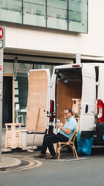 A man with glasses, wearing a light blue shirt and dark trousers, is seated on a wooden chair outside a white commercial van on a city street during daytime. The van's side door is open, revealing an interior filled with cardboard boxes, wooden furniture panels, and packing materials. Moving blankets are draped inside, and a wooden stand is positioned in front of the van. The scene shows objects typically used in home relocation and furniture transport, with the man appearing to take a break amidst the loading or unloading process. The background features a modern building with large glass windows and a grey facade, indicating an urban environment, while the pavement and street markings are also visible. This image exemplifies house removals and packing activities, relevant to services provided by Man with Van Brixton.