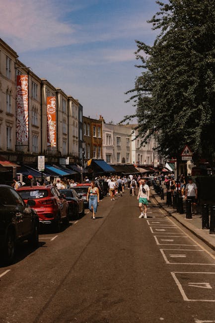 An aerial view of a busy street in Brixton, showing multiple vehicles including cars, vans, and buses moving along the road, with some vehicles parked along the side of the pavement. On the left side, there are commercial buildings and a sidewalk with pedestrians walking, while on the right side, there are residential houses and more pedestrians. The street is lined with green trees providing shade, and in the background, a variety of urban structures are visible, including multi-story buildings and rooftops. The scene captures the typical urban environment relevant to house removals and moving services, highlighting the logistics of furniture transport and home relocation, possibly involving companies like Man with Van Brixton, who manage loading processes and transportation logistics within a busy residential and commercial area.
